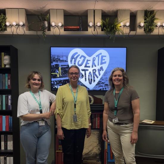 Matilde, Nina og Helle står smilende sammen på Lyrikscenen på Hovedbiblioteket. Bag dem ses en skærm med reklame for podcasten "Hjertestorm". Over dem ses en lyskæde.