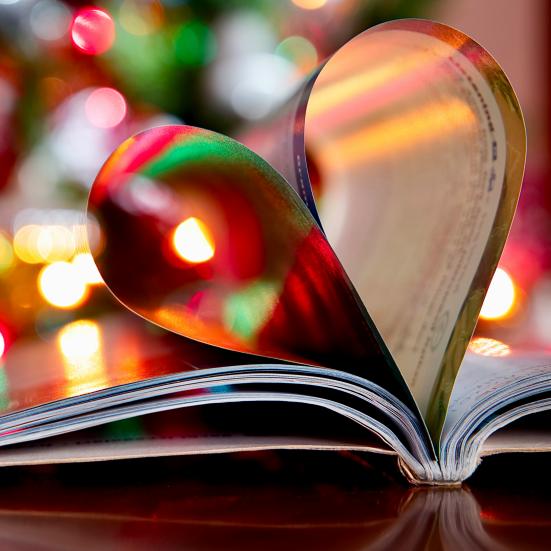 En bog ligger opslået, dens sider danner et hjerte. I baggrunden anes julelys, baggrunden er sløret.