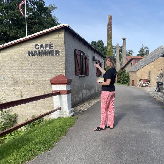 Line står uden for Café Hammer i Svenstrup. Hun holder armen frem mod bygningen og ser glad ud. I den anden arm har hun en stak bøger.