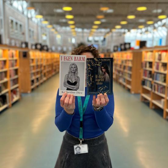 Ditte står i det store udlån på Hovedbiblioteket. Hun holder bøgerne "I egen barm" af Katherine Diez og "I den mørke skov" af Gry Kappel Jensen op foran sit ansigt og strakt frem for sig. Baggrunden er let sløret, men man fornemmer tydeligt biblioteksrummet med de mange fyldte bogreoler. 