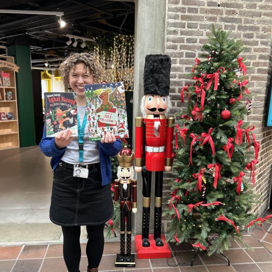 Ditte står foran indgangen til børnebiblioteket på Hovedbiblioteket. Ved siden af hende ses juletræ og julepynt. Hun holder bøgerne "Iqbal Farooq og julesvineriet" og "Hvor er julepøllerne?" frem for sig, mens hun smiler.