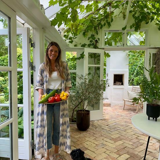 Foto af Maria Lundvald i en udestue. Der er frodigt grønt i loftet, og man kan også se den grønne have udenfor gennem vinduerne. Maria smiler og holder et fad med grøntsager. Ved hendes fødder sidder en lille hund. 