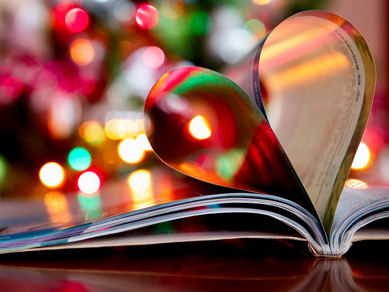 En bog ligger opslået, dens sider danner et hjerte. I baggrunden anes julelys, baggrunden er sløret.