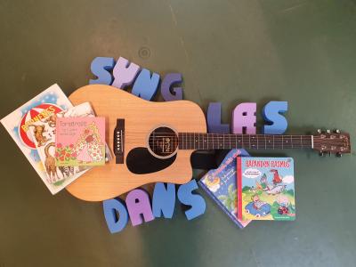 En guitar ligger på et gulv. Der ligger billedbøger på og omkring guitaren, og skumbogstaver, der danner ordene "syng", "læs" og "dans"