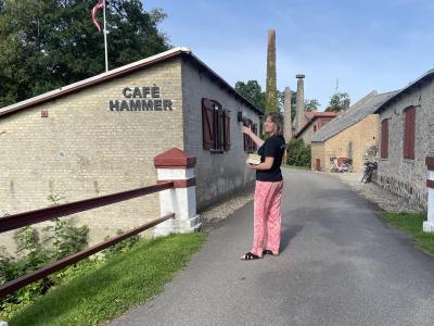 Line står uden for Café Hammer i Svenstrup. Hun holder armen frem mod bygningen og ser glad ud. I den anden arm har hun en stak bøger.