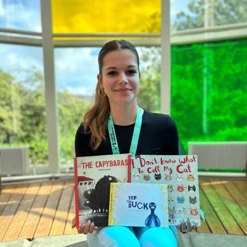 Nina sidder smilende i Glashjørnet på Hovedbiblioteket. Hun holder de tre billedbøger, som hun anbefaler i artiklen. Bag Nina ses Glashjørnets farverige ruder.