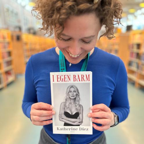 Ditte står med bogen "I egen barm" af Katherine Diez. Hun holder bogen op foran sig, så man kan se bogens forside. Ditte smiler og kigger ned på bogen. Baggrunden er sløret, men man fornemmer biblioteksrummet med de mange fyldte bogreoler.