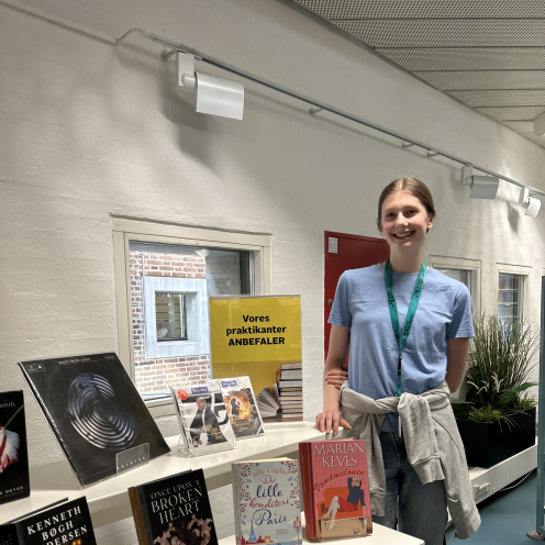 Frederikke står ved siden af sin udstilling på Hovedbiblioteket. På udstillingen ses forskellige bøger, film og musik. Frederikke smiler til kameraet.