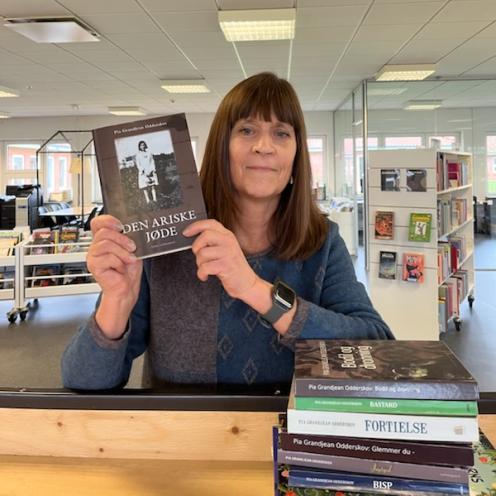 Karin står på biblioteket med et udvalg af Pia Grandjeans bøger stående i en stak foran sig. Hun holder "Den ariske jøde" op. Bag Karin ses biblioteksrummet.