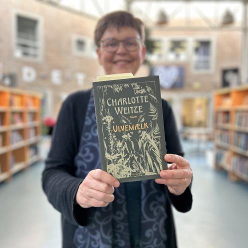 Vita står på Hovedbiblioteket. Hun holder bogen "Ulvemælk" frem foran sig, så der er fuld fokus på forsiden. Både Vita og baggrunden er let sløret, men man kan se, hun smiler og at hun er omgivet af fyldte bogreoler.