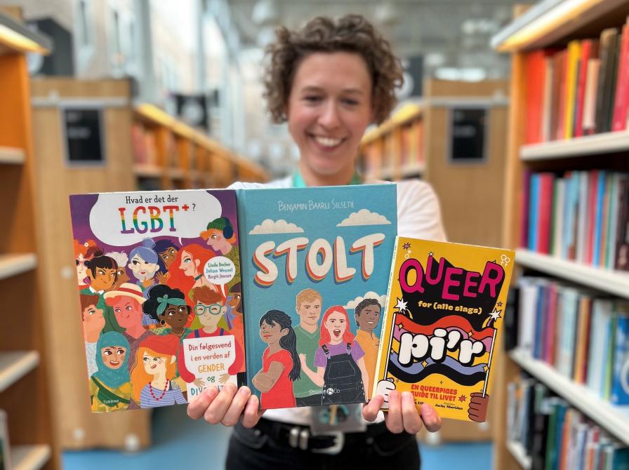 Ditte står smilende blandt fyldte bogreoler på Hovedbiblioteket. Frem for sig holder hun tre bøger med LGBT+-tema. Bogforsiderne er i fokus, mens baggrunden og resten af Ditte er let sløret.