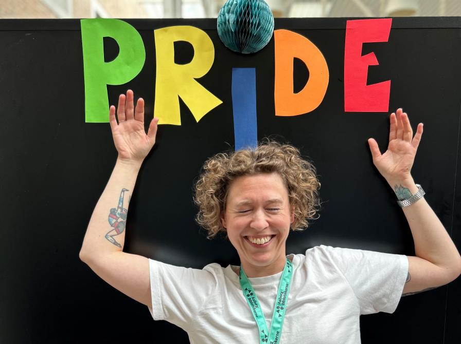 Ditte står foran en sort baggrund, hvorpå der står "Pride" i forskellige farver over hendes hoved. Ditte smiler stort, kniber øjnene sammen og har løftet sine arme op mod teksten med "Pride".