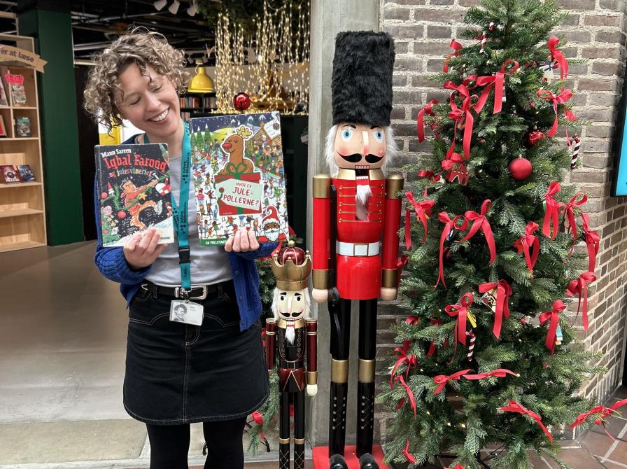 Ditte står foran indgangen til børnebiblioteket på Hovedbiblioteket. Ved siden af hende ses juletræ og julepynt. Hun holder bøgerne "Iqbal Farooq og julesvineriet" og "Hvor er julepøllerne?" frem for sig, mens hun kigger ned på dem og smiler.