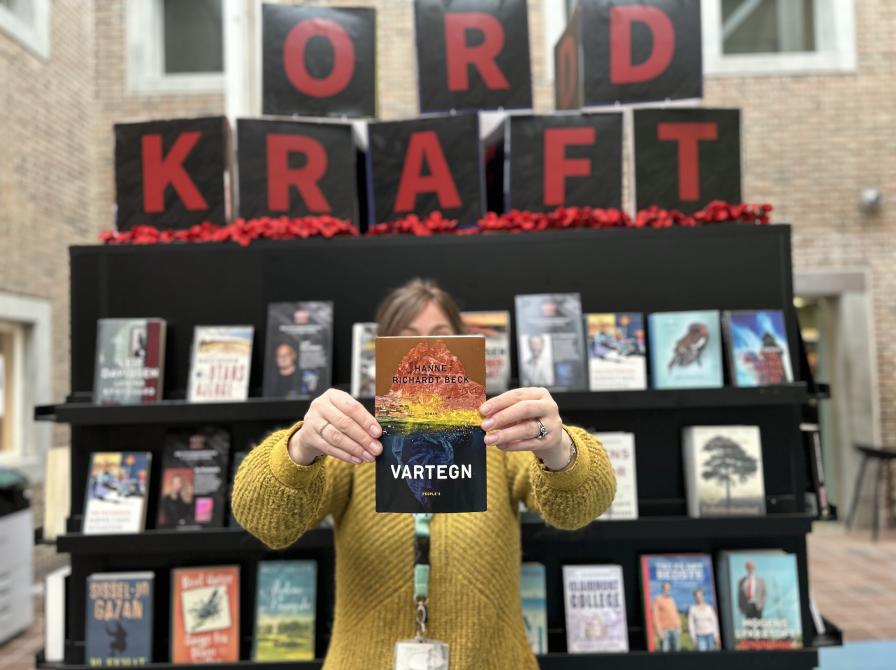 Line står på Hovedbiblioteket foran Ordkraft-udstillingen, som er fyldt med bøger af forfattere, der gæster festivalen i 2025. Udstillingen er øverst dekoreret med klodser, som danner ordet ORDKRAFT. Line holder "Vartegn" frem for sig i strakte arme, så den dækker hendes ansigt. Både Line, udstillingen og resten af biblioteksrummet er let sløret, så der er fokus på bogen i hendes hænder.