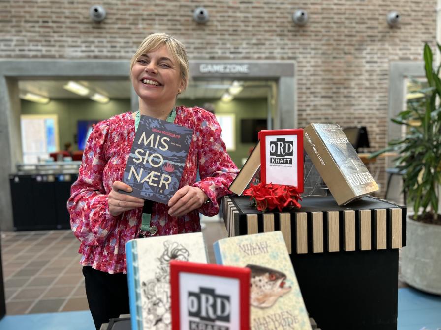 Marianne står ved Ordkraft-udstillingen på Hovedbiblioteket, hvor der er udstillet bøger af forfattere, der gæster festivalen i går. Marianne smiler og holder "Missionær" i hænderne. Bag hende ses biblioteksrummet, som er let sløret.