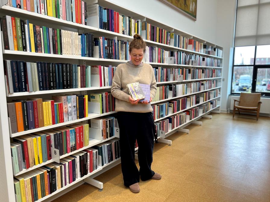 Michelle står på biblioteket. Hun holder "Øvelser i mørke" og "Poesibog" i hænderne og kigger smilende ned på bøgerne. Bag Michelle ses fyldte bogreoler.