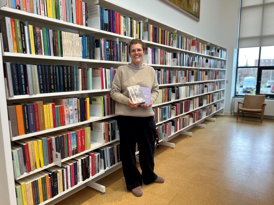 Michelle står på biblioteket. Hun holder "Øvelser i mørke" og "Poesibog" i hænderne og kigger smilende ind i kameraet. Bag Michelle ses fyldte bogreoler.