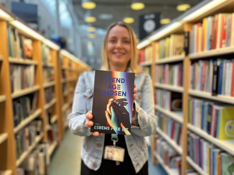 Nina står på Hovedbiblioteket med fyldte bogreoler til hver side af sig. Hun smiler og holder bogen "Esbens fire liv" frem for sig. Der er fokus på bogforsiden, mens både Nina og resten af biblioteksrummet er let sløret.